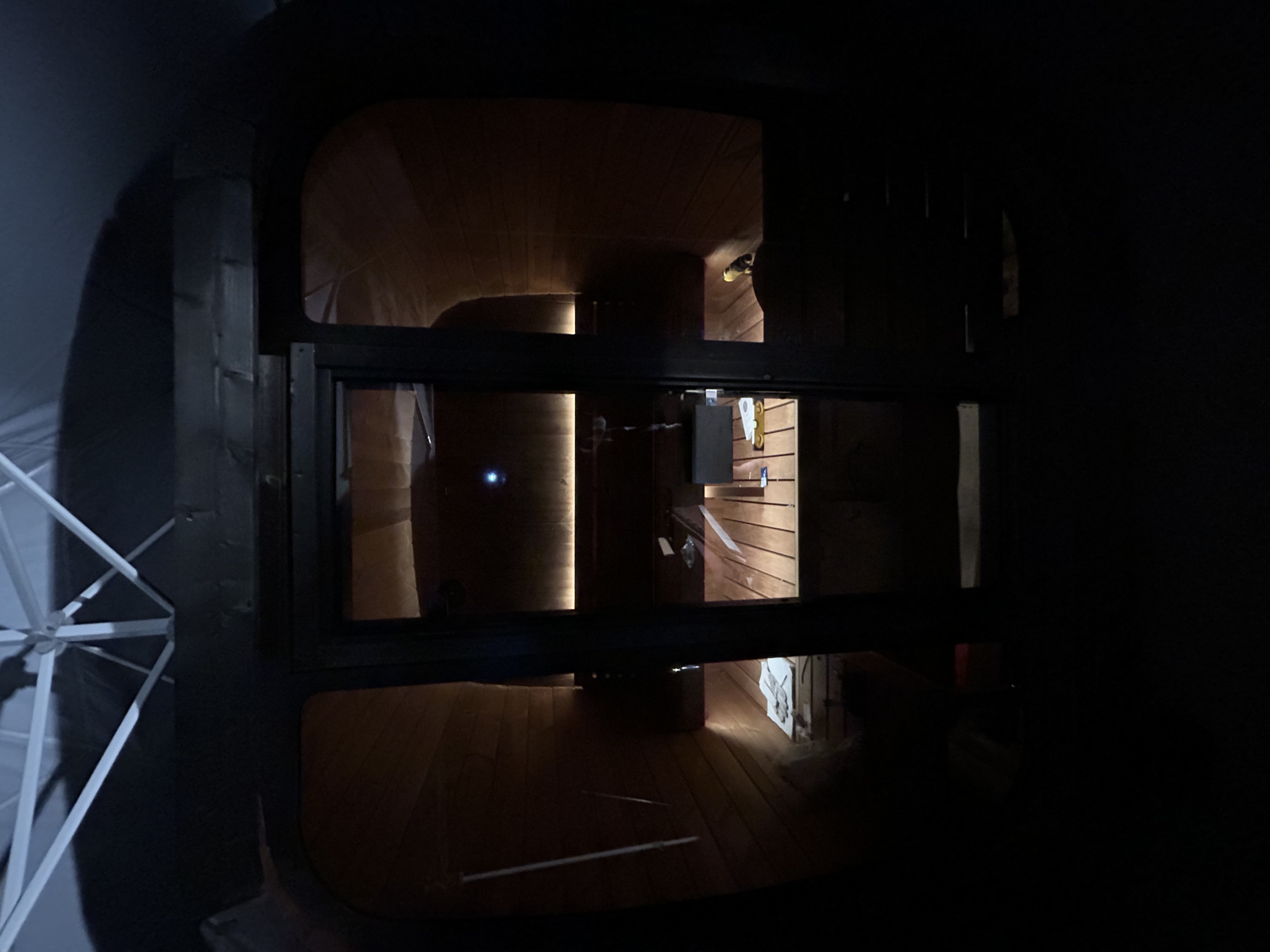 Dark exterior view looking through a glass door into a wooden sauna lit by warm LED backrest lights.